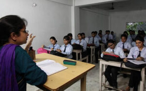 Classroom scene with a teacher instructing a group of students in formal uniforms, taking notes at desks. The classroom has plain white walls and natural light coming through the windows.